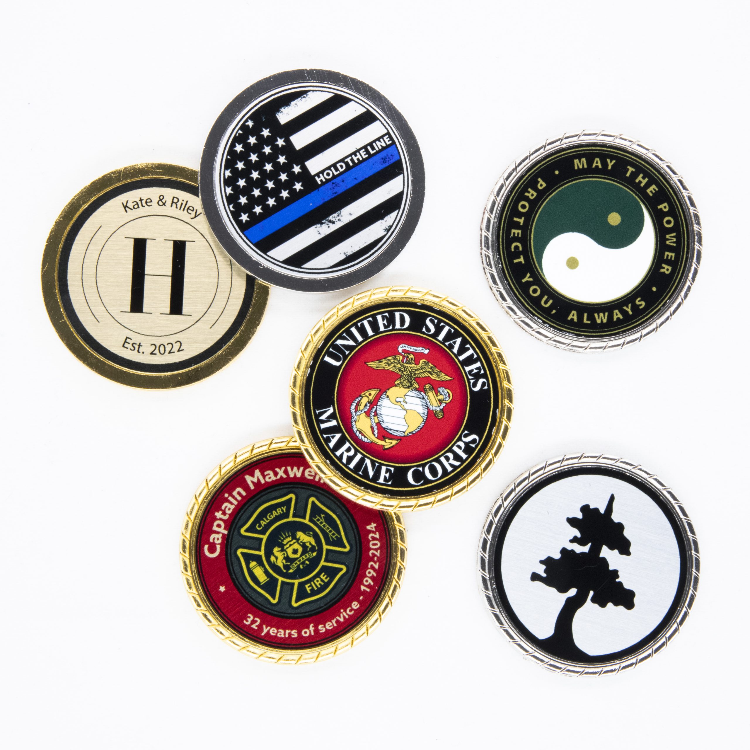 Assortment of personalized challenge coins displayed on a white background. Designs include a United States Marine Corps coin with the eagle, globe, and anchor emblem, a 'Hold the Line' thin blue line flag coin, a Yin-Yang coin with 'Protect You, Always' text, a custom firefighter coin for 'Captain Maxwell' from Calgary with 32 years of service, a personalized coin with 'H' and 'Kate & Riley' and 'Est. 2022', and a black and white coin featuring a tree or nature silhouette
